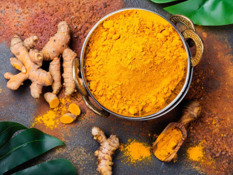 Sveži koren kurkume u drvenoj činiji koja stoji na podlozi od kurkume u prahu.