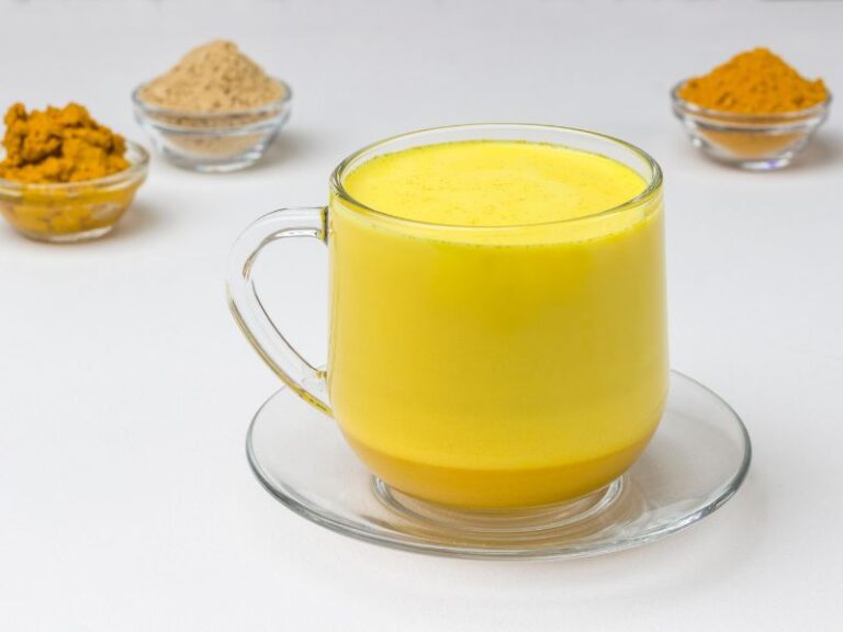 Zlatno mleko sa kurkumom, đumbirom i cimetom, prikazana na beloj pozadini sa činijicama začina u pozadini.