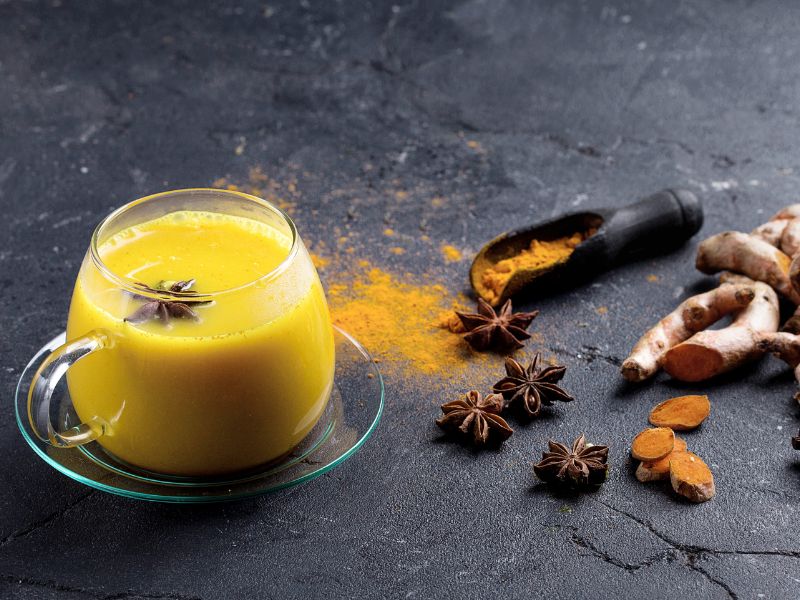 Šolja zlatnog mleka sa anisom na tamnoj podlozi, okružena kurkumom u prahu i svežim korenom kurkume